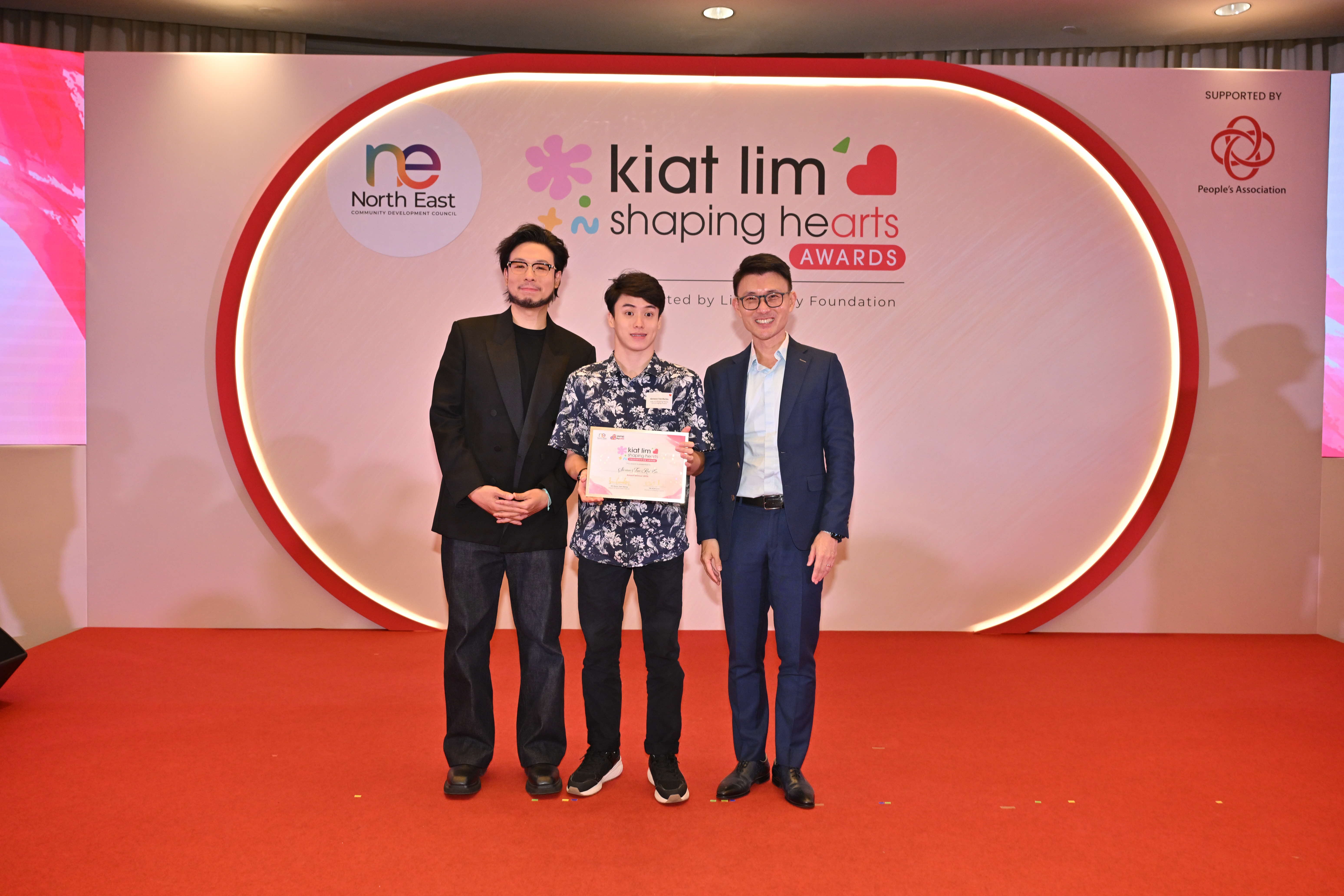 Award recipient standing on stage with the presenter, holding their certificate and posing for a group photo during the award ceremony, with a backdrop reading ‘Kiat Lim-Shaping Hearts Awards'.